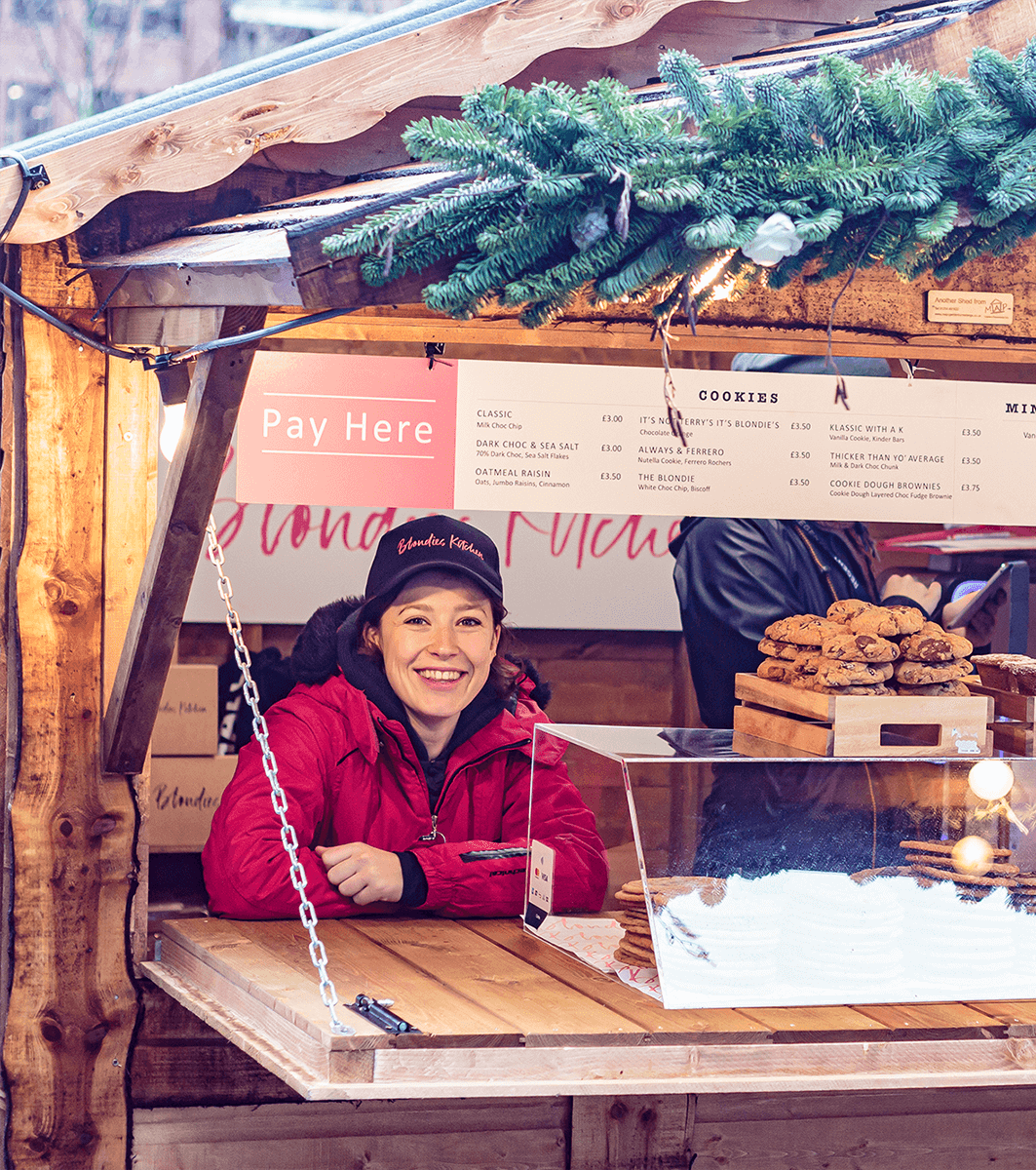 Brunette female brand ambassador wearing a baseball cap working at a Blondies Kitchen pop-up kiosk at London market. There is a selection of cookies on the counter beside her. 