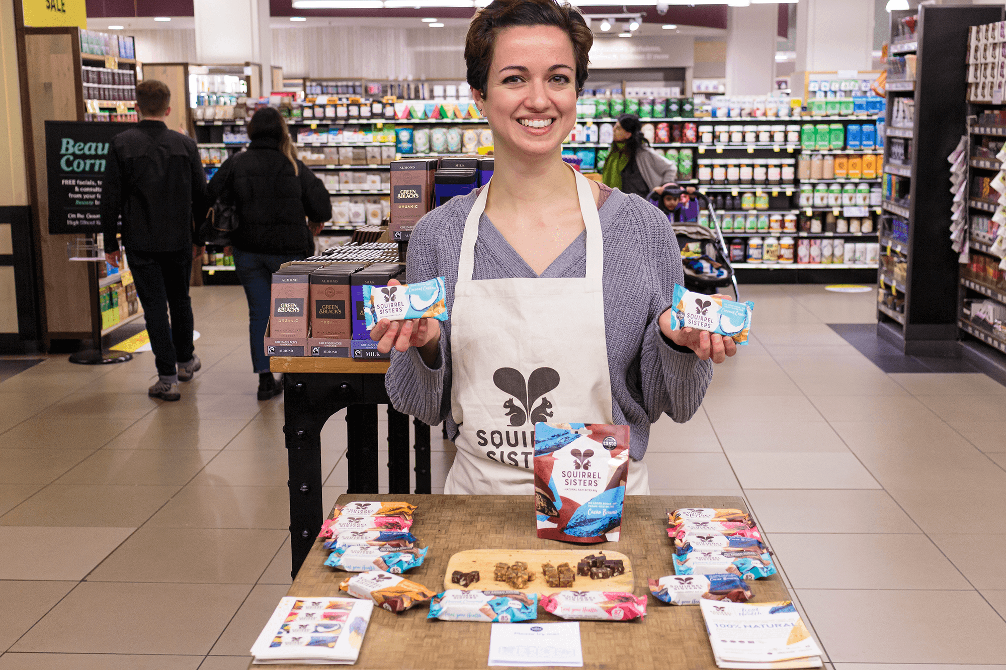 Female brand ambassador with short, dark hair standing behind a table giving out free samples of Squirrel Sisters healthy snack bars.