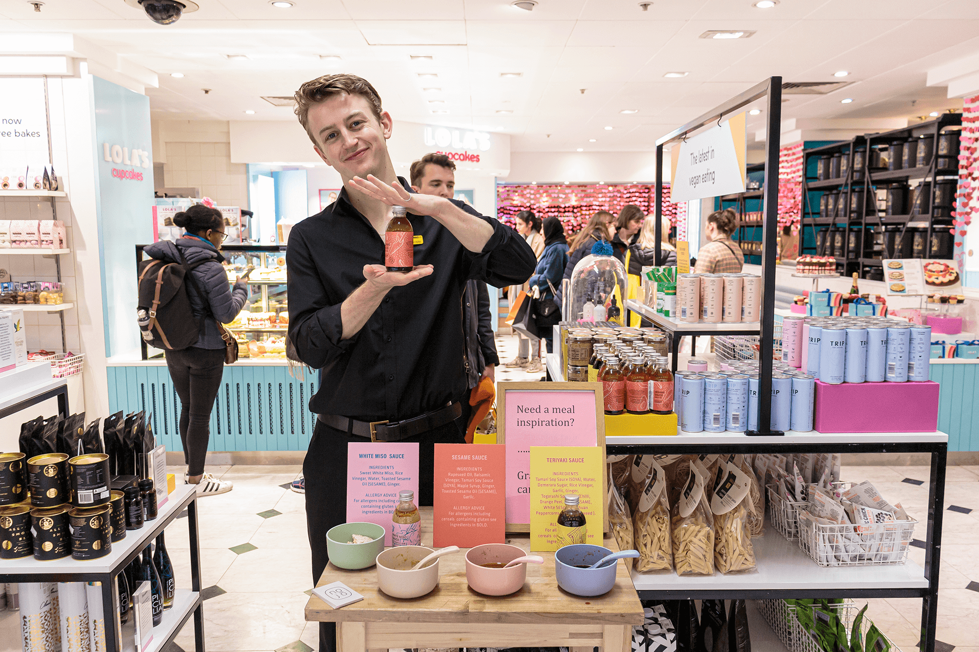 Young man with blonde hair wearing a black shirt. He is standing behind a small wooden table at Selfridges, Oxford street in the Food Hall. He is holding a small bottle of Noju Japanese  marinade. 