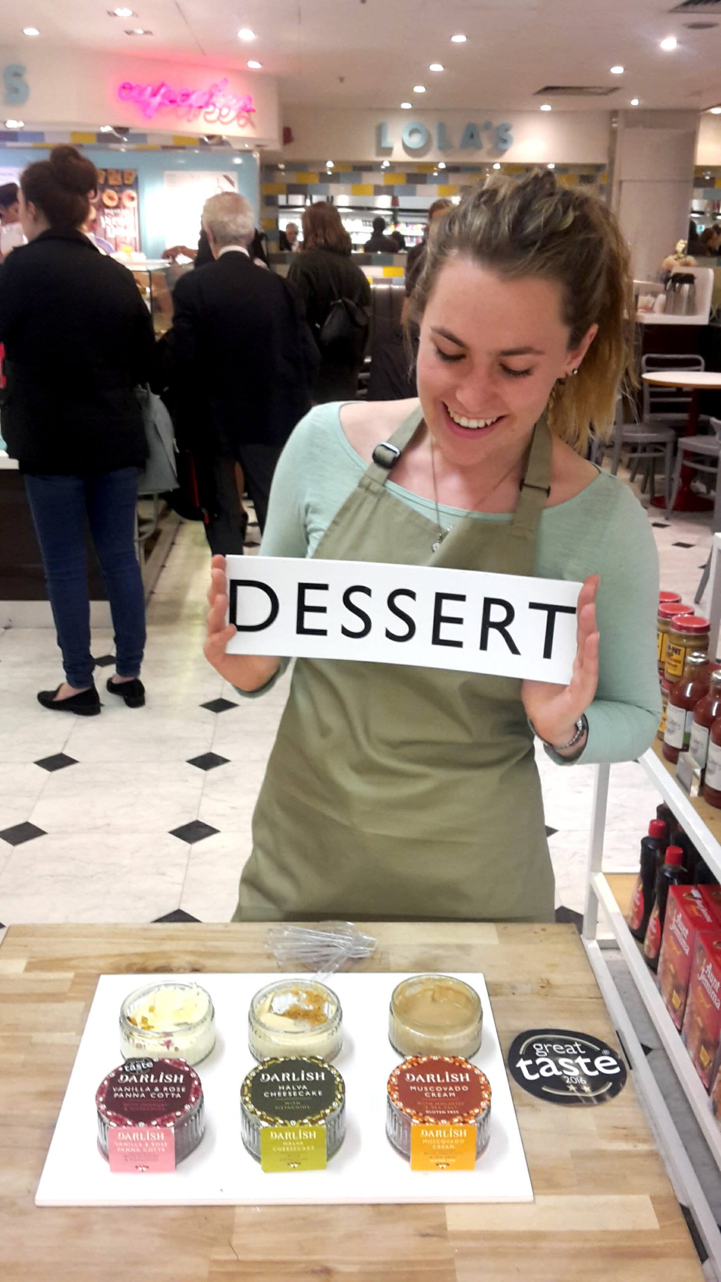 Young blonde woman looking downwards at a black and white ‘dessert’ sign that she is holding. She is standing behind a table with dessert pots laid out on it. 