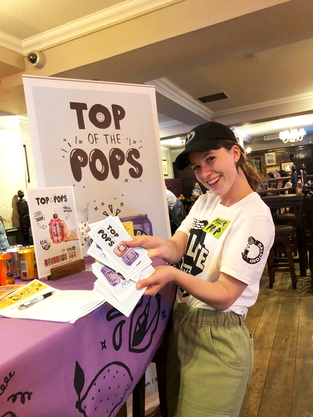 Young woman smiling and wearing a baseball cap and Dalston’s Soda branded t-shirt. She is fanning some promotional leaflets in front of a table with a purple tablecloth and a marketing banner.  