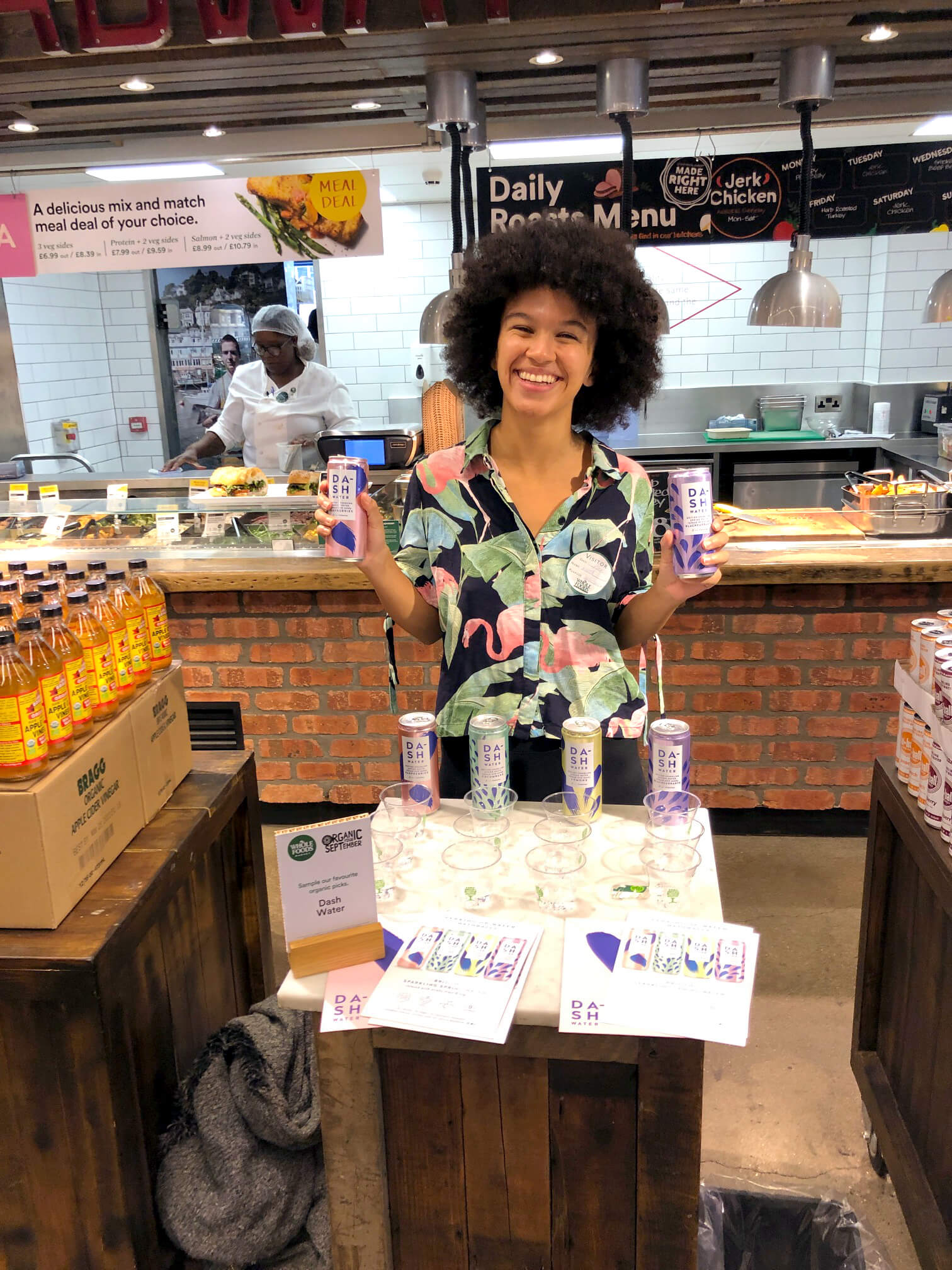 Mixed-race young woman with afro hair smiling whilst standing behind a table. She is holding 2 cans of ‘Dash’ sparkling water.