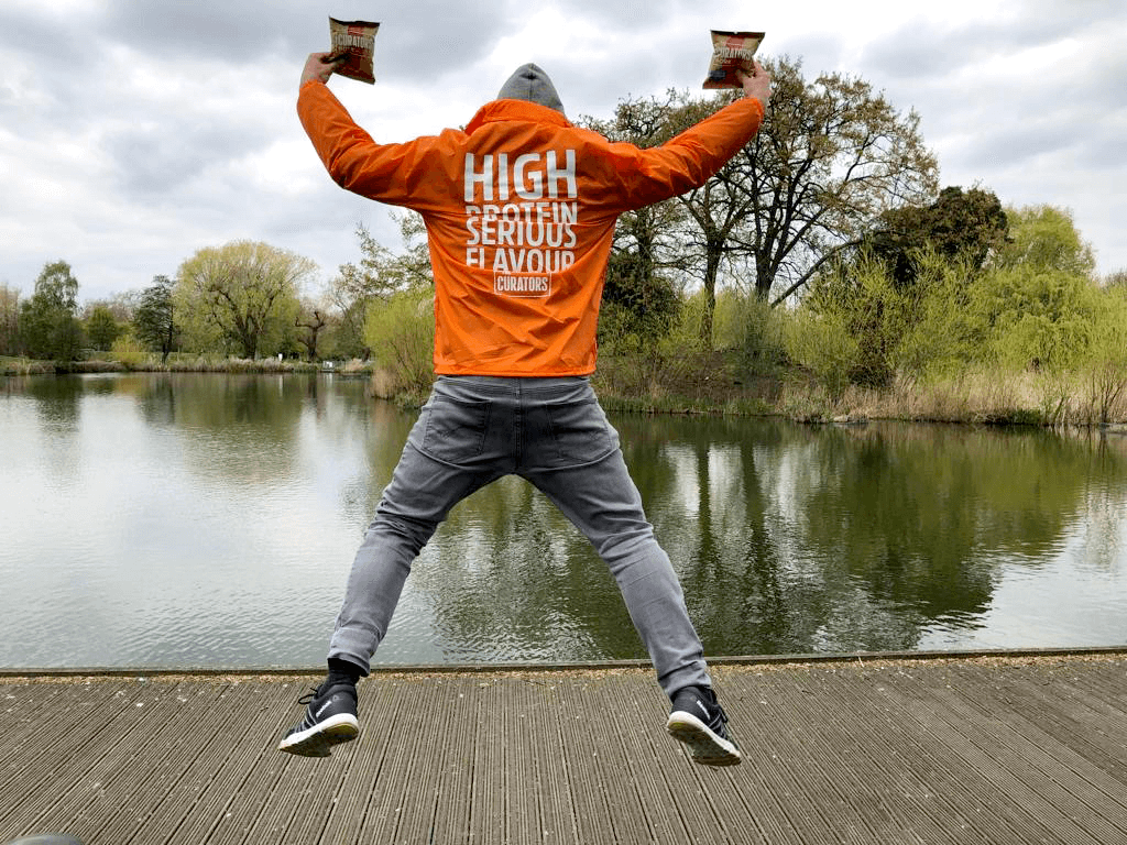 Male with his back facing the camera jumping in the air in a star-jump position. He is wearing an orange waterproof jacket with the words ‘High Protein, Serious Flavour, The Curators’ on it and holding 2 packets of ‘The Curators’ Pork puff snacks. 