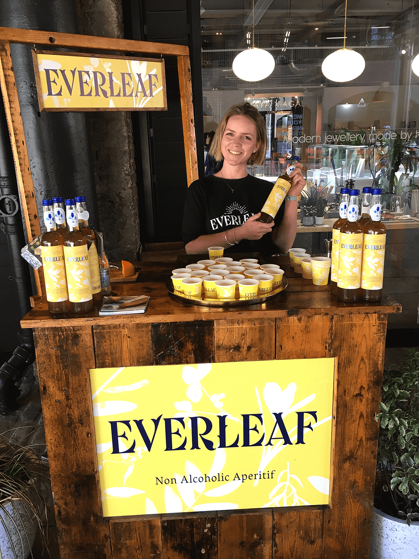 Female brand ambassador with a blonde bob is standing behind a table at an event pouring drinks of Everleaf alcohol-free spirit. 