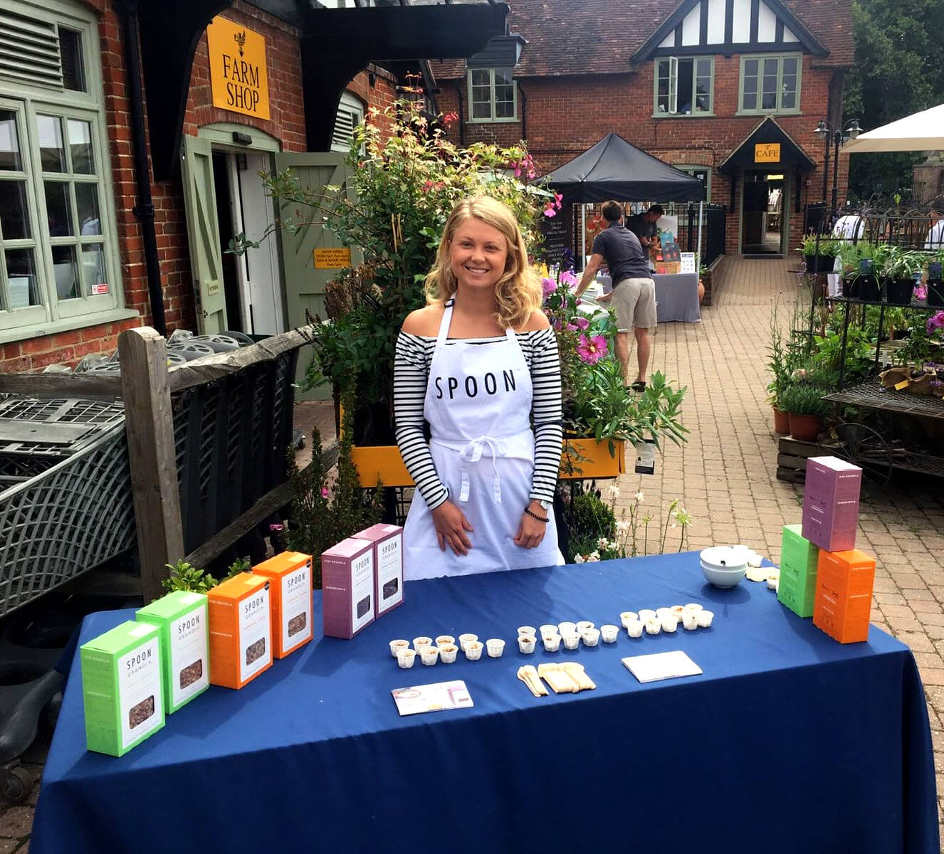 Blonde woman smiling and wearing a white apron with ‘Spoon’ on it. She is standing behind a blue table with ‘Spoon’ cereal boxes and some sample pots and leaflets on it.  