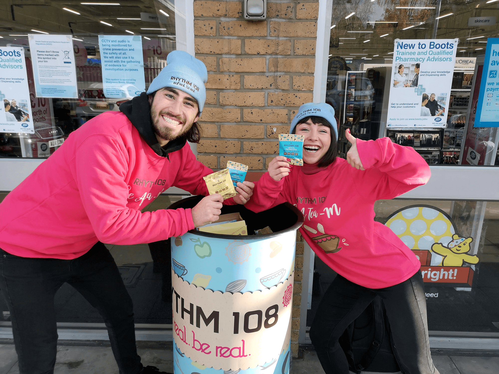 Male and Female brand ambassadors standing outside of Boots store High Holborn. They have a Rhythm108 branded wheelie bin and are giving out free samples of Rhythm108 gluten-free, vegan biscuits to members of the public. 